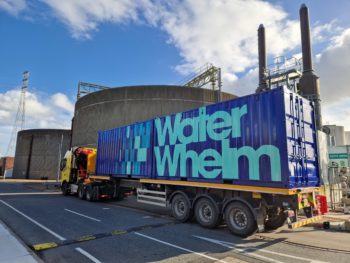 An image of Waterwhelm's desalination pilot plant in a bright blue shipping container, with the words 'Waterwhelm' on the side, placed on the back of a lorry on a road.