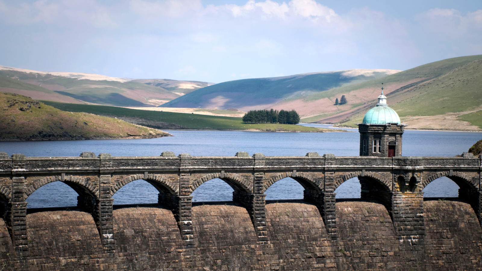 The Old Dam on the River Elan, in Wales. Behind the dark brick dam wall is a still lake, in front of green hills.