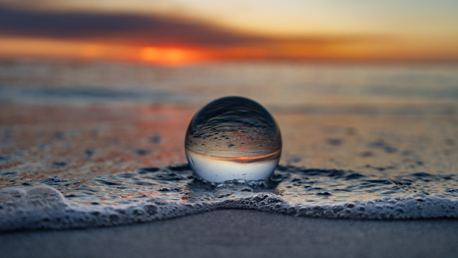 A clear glass globe on the beach at sunrise. The sea foam and orange sky are reflected upside down within the globe.