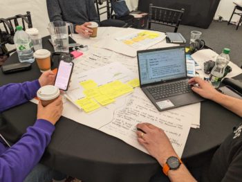 Image of three people sat at a table, with a laptop and big sheets of paper and post-it notes with writing.