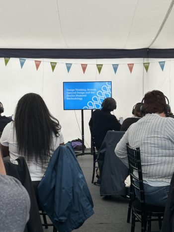 Image of a group of people seated around tables, looking at TV screen. Text on screen says 'Design Thinking, Human Centred Design and the Double Diamond Methodology' against a blue background.