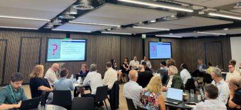 Image of attendees at the Water Discovery challenge final event, with attendees sat around tables and listening to four speakers at the front of the room sharing their insights in a 'fishbowl' format