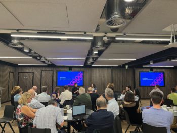 Image of attendees at the Water Discovery Challenge Final Event, sat around tables and listening to a conversation with keynote speaker, Kenny Ewan. Kenny is sat at the front of the room alongside Caroline, who is asking questions.