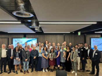 An image of almost 40 attendees of the Water Discovery Challenge final event, standing at the back of a wood panelled room in front of screens, facing and smiling at the camera