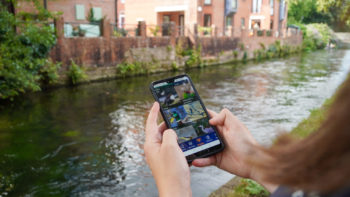 A point-of-view shot of someone using the 'Big River Watch app' in front of a canal. The app shows pictures of wildlife which may be visible near a canal, such as ducks, fish and herons.