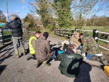 Image of six people wearing outdoor gear in a road surrounded by fields and trees on a sunny day. Five people are sat on the floor around laptops and talking. One person is stood up facing away holding a test tube between gloved hands.